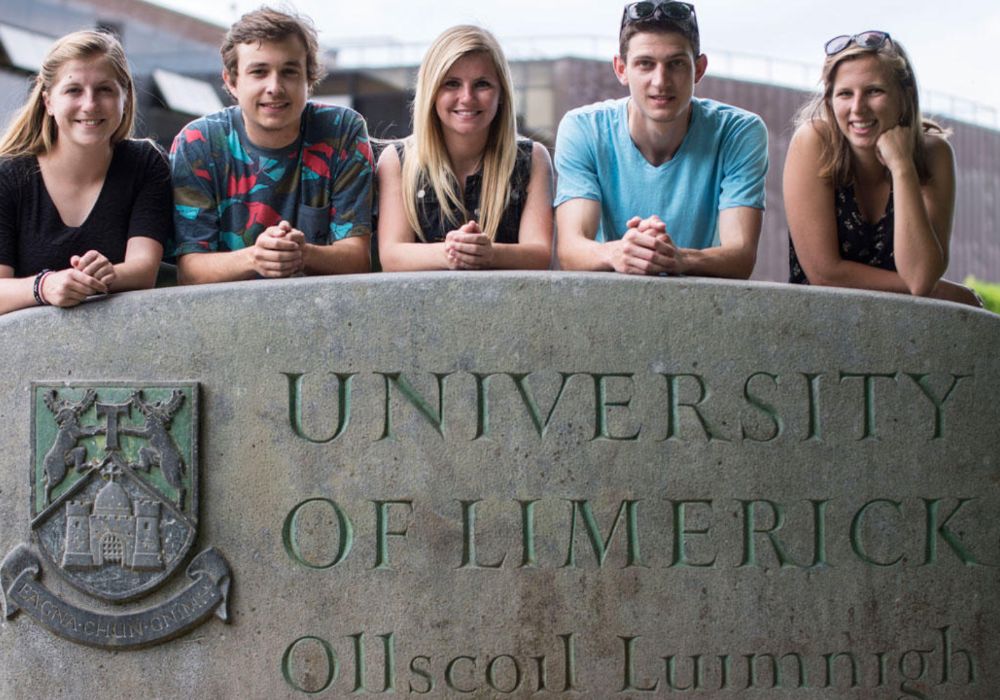 A group of young people smiling and posing in front of the University of Limerick sign.